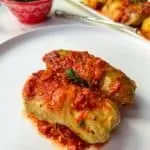 two stuffed cabbage rolls with tomato sauce on a white plate, with a white serving plate with stuffed cabbage rolls and a red bowl filled with parsley in the background
