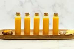 lemon ginger turmeric wellness shots recipe in tiny glass vials on a wooden plate/surface with a piece of ginger and turmeric root on each side